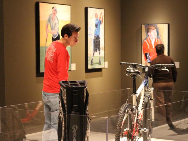 A photo of a visitor looking at a display featuring a Trek bicycle and a gold bag, part of the 2017 exhibit Portraits of Courage, at the George W Bush Library and Museum